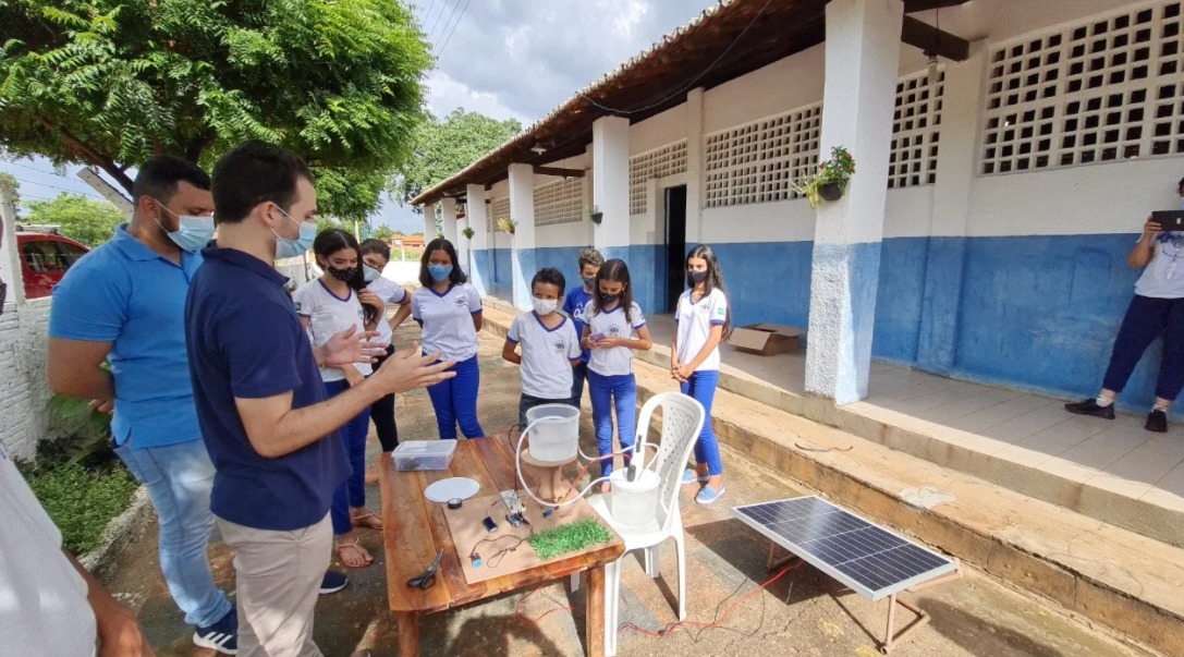 professor dando aula em frente a uma mesa com objetos do experimento solar e alunos observando com escola branca e azul ao fundo